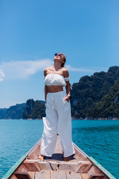 Fashion portrait of young woman in white top and pants on vacation, on sailing thai wooden boat. Travel concept. Female in Khao Sok National park.