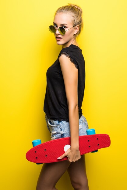 Fashion pretty cool girl in sunglasses with skateboard over colorful yellow background