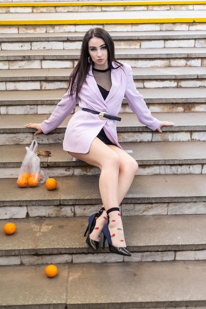 과일 오렌지가 있는 패션 예쁜 여성 모델은 보라색 재킷 검은색 신발을 신고 계단에 앉아 있습니다. 프리미엄 사진