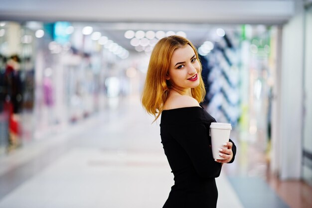 Fashion red haired girl wear on black dress with bright make holding coffee cup at trade shopping center Photo toned style Instagram filters