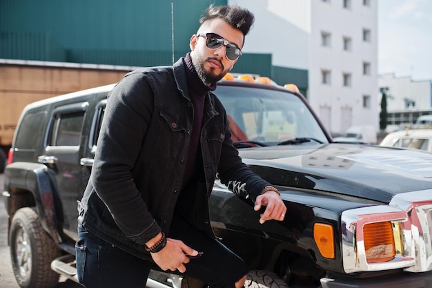 Foto gratuita moda ricca barba uomo arabo indossare su giacca di jeans neri e occhiali da sole in posa contro una grande automobile nera suv elegante modello arabo di successo e alla moda