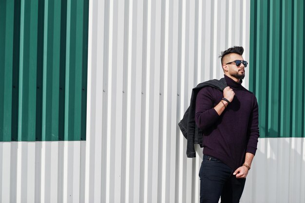 Fashion rich beard Arab man wear on turtle neck and sunglasses posed against green and white steel wall with jacket on hand Stylish succesful and fashionable arabian model guy