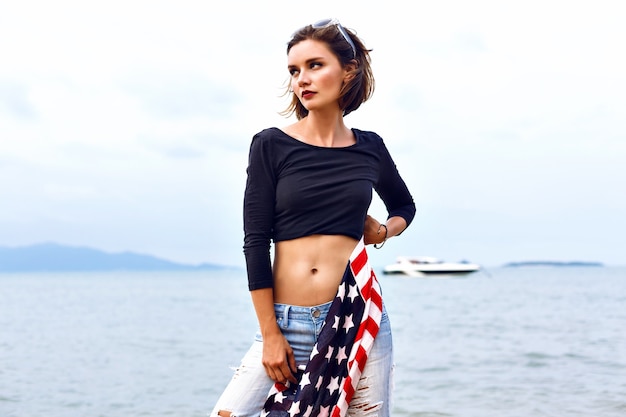 Fashion sensual portrait of beautiful sad woman posing on the island beach at windy rainy day, wearing old school  outfit, have fit sexy bode, holding american flag in her hands.