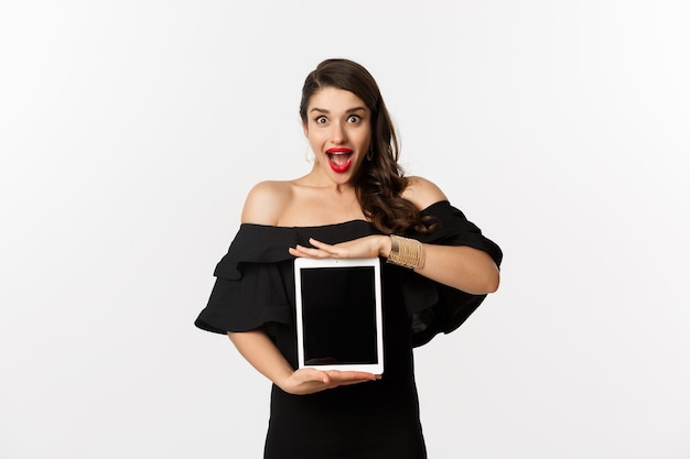 ファッションとショッピングのコンセプト。タブレット画面でオンラインウェブサイトのプロモーションオファーを表示し、興奮したカメラを見て、黒いドレス、白い背景で立っている驚いた若い女性。