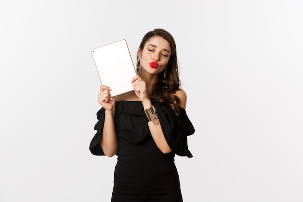 Fashion and shopping concept. beautiful woman with red lips, wearing black dress, showing digital tablet and making kissing face, white background