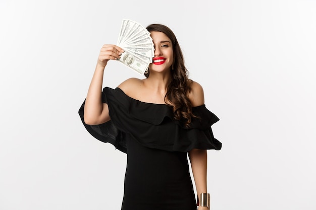 패션과 쇼핑 개념. 행복 한 젊은 여자 검은 드레스, 붉은 입술, 돈을 들고 웃 고 만족, 흰색 배경 위에 서 서.