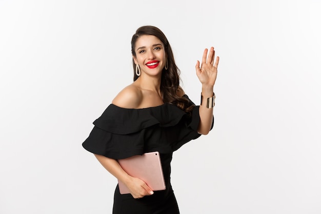 Fashion and shopping concept. Stylish young woman with glamour makeup, wearing black dress, holding digital tablet and saying hi, waving hand to greet you, white background.