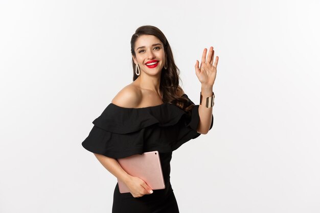 Fashion and shopping concept. Stylish young woman with glamour makeup, wearing black dress, holding digital tablet and saying hi, waving hand to greet you, white background