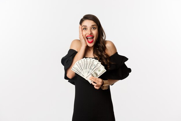 Fashion and shopping concept.  woman rejoicing of money prize, holding dollars and shouting from excitement, standing in black dress over white background.