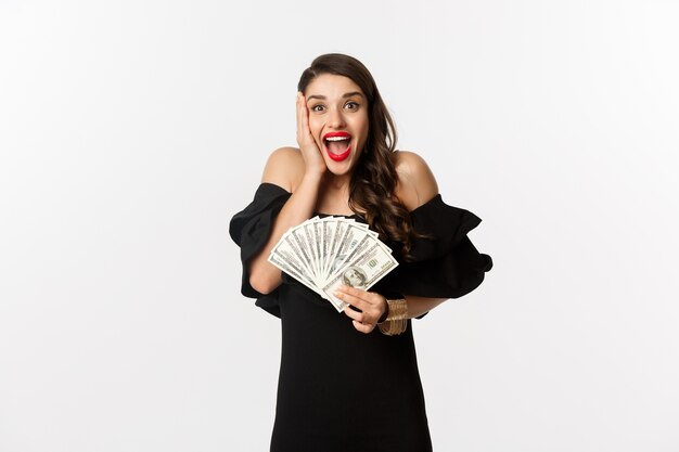 Концепция моды и покупок. женщина, радуясь денежной премии, держа доллары и крича от волнения, стоя в черном платье на белом фоне.