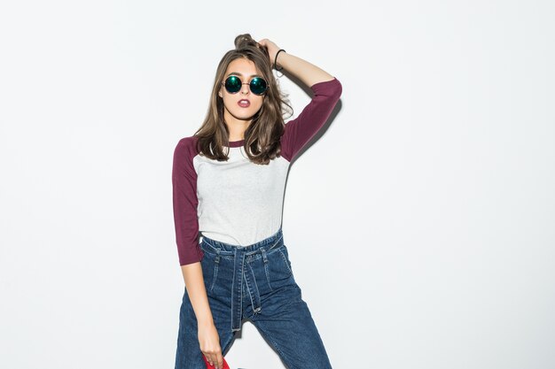 Fashion skater girl in casual clothes and black sunglasses holding red skateboard isolated on white wall