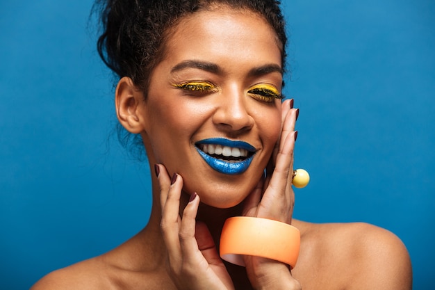 Fashion smiling mulatto woman with colorful makeup and curly hair in bun touching her pretty face and looking on camera isolated, over blue wall