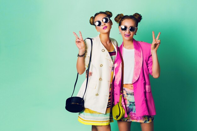 세련 된 캐주얼 봄 복장 재미, 쇼 혀에 두 젊은 여성의 패션 스튜디오 이미지. 밝은 유행 색상, 만두가있는 세련된 헤어 스타일, 멋진 선글라스. 친구 초상화.
