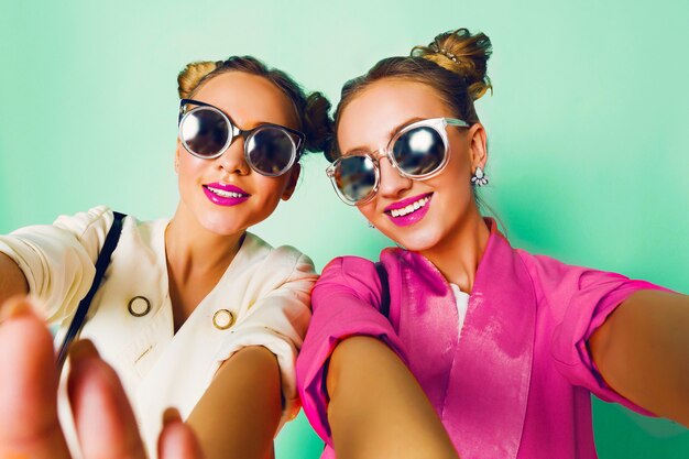 세련 된 캐주얼 봄 복장 재미, 쇼 혀에 두 젊은 여성의 패션 스튜디오 이미지. 밝고 트렌디 한 파스텔 색상, 만두가있는 세련된 헤어 스타일, 멋진 선글라스. 친구 초상화.