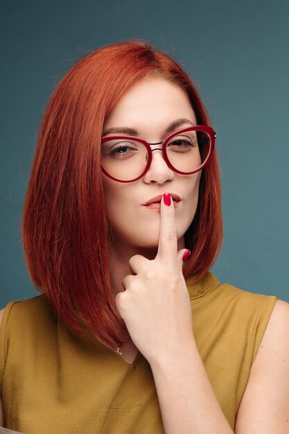 Модный студийный портрет симпатичной молодой хипстерской рыжеволосой женщины с ярким сексуальным макияжем и очками в стильной городской футболке Синий фон стены
