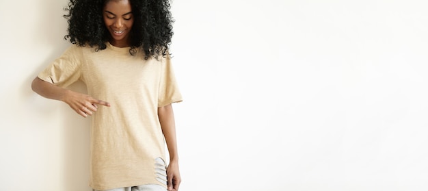 Fashion, style, design and clothing concept. Cropped shot of good-looking young African woman with Afro haircut wearing stylish ripped oversize t-shirt, pointing finger at it and smiling joyfully