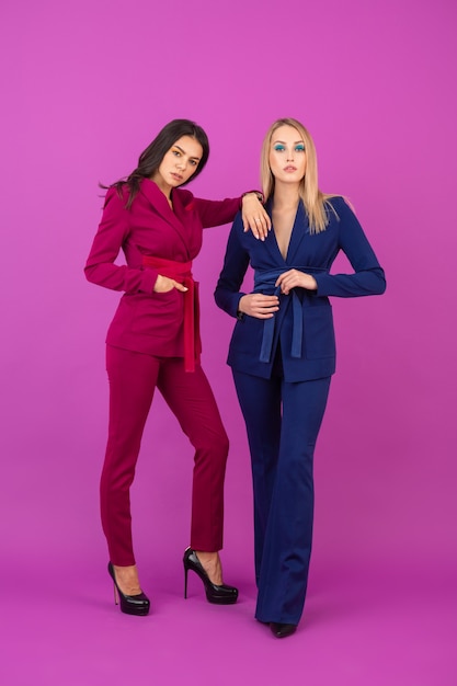 Модный стиль две улыбающиеся привлекательные женщины на фиолетовой стене в стильных красочных вечерних костюмах фиолетового и синего цвета, весенняя модная тенденция