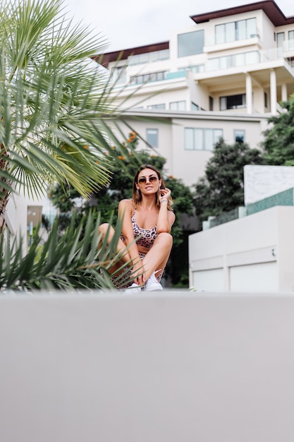 Foto gratuita moda donna europea abbronzata vestibilità elegante in occhiali da sole, canotta leopardata e pantaloncini da motociclista, fuori dalla villa vicino alla palma