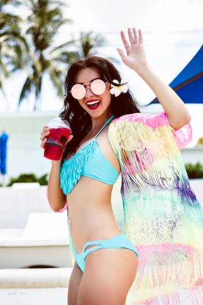 おいしいレモネードを飲んで見事な官能的な日焼けした女性のファッション夏の肖像画。トレンディなビーチウェアとサングラスを身に着けている高級ホテルで彼女の休暇を楽しんでください。