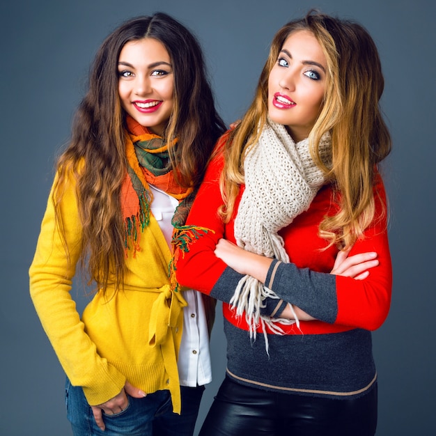 Fashion winter portrait of blonde and brunette beautiful best friends girls, hugs and having fun. Wearing bright stylish cashmere sweaters and scarfs. Have trendy makeup and long amazing hairs.