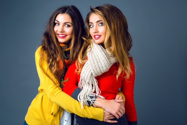 Fashion winter portrait of blonde and brunette beautiful best friends girls, hugs and having fun. Wearing bright stylish cashmere sweaters and scarfs. Have trendy makeup and long amazing hairs.