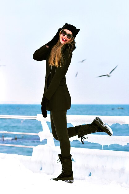 エレガントなブロンドの女性の冬のポートレートをファッションし、海で冷たい雪の時間を楽しみ、氷と風が強く、黒いコート、変な帽子、長い髪、官能的な気分、一人旅、冬のファッション。