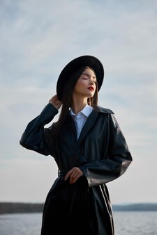 호수 연못 근처에 검은색 둥근 모자와 가죽 비옷을 입은 패션 여성. 해질녘 초상화 소녀입니다. 입술에 아름다운 메이크업과 밝은 빨간 립스틱