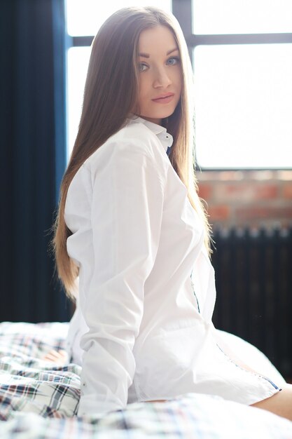 Fashion young woman posing with male shirt, sitting in the bed