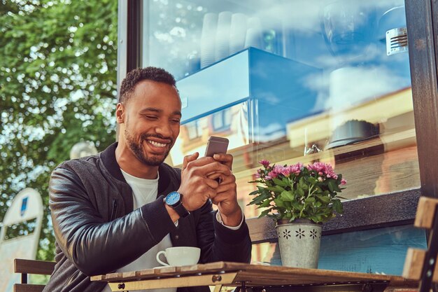 A fashionable African-American bearded male sitting near a coffee shop with a cup of coffee, using a smartphone.