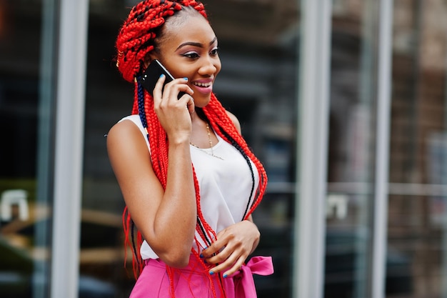 Fashionable african american girl at pink pants and red dreads posed outdoor with mobile phone
