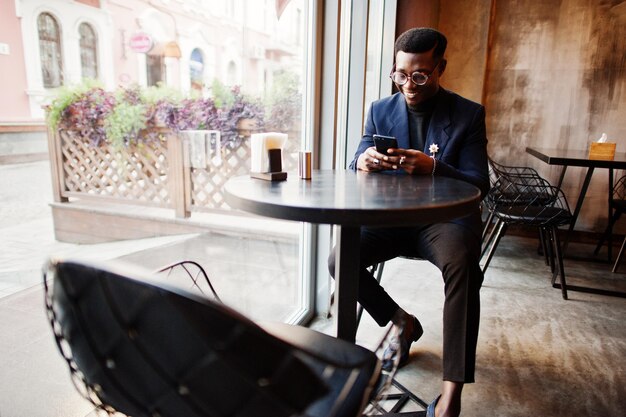 Модный афроамериканец в костюме и очках, держащий мобильный телефон в руках, позирует в кафе