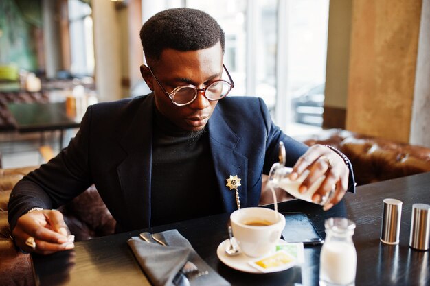 Fashionable african american man in suit and glasses pours cream into coffee at cafe