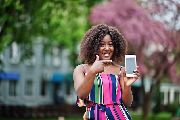 Модная африканская американка в розовом полосатом комбинезоне позирует на улице весеннего цветения и показывает пальцем на экран мобильного телефона