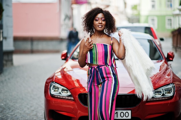 Fashionable african american woman in pink striped jumpsuit with fluffy faux fur coat posed against rich red luxury car with mobile phone at hand