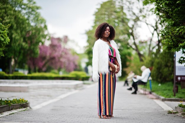 Fashionable african american woman in pink striped jumpsuit with fluffy faux fur coat posed at spring bloom street