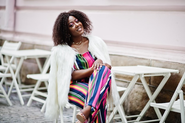 Fashionable african american woman in pink striped jumpsuit with fluffy faux fur coat sitting on chair at outdoor cafe