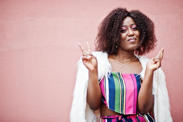 Free photo fashionable african american woman in striped jumpsuit with fluffy faux fur coat and handbag posed against pink wall