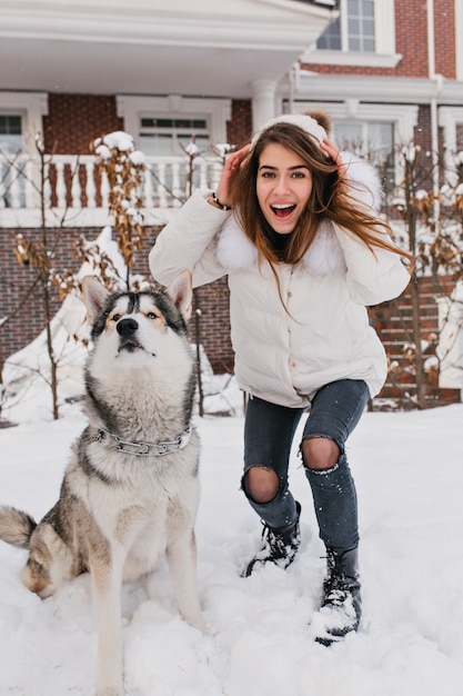 雪の中で屋外のかわいいハスキー犬を楽しんでファッショナブルな素晴らしい女性。本物の友達、家庭のペット、愛する動物の幸せな冬時間