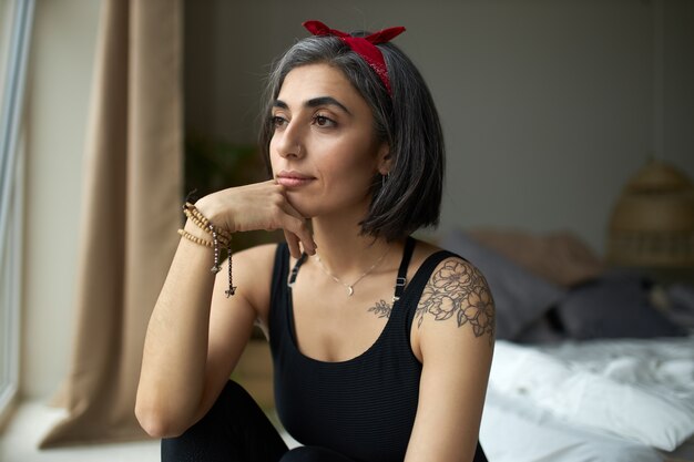 Fashionable attractive tattooed young woman sitting in bedroom by window with pensive thoughtful look