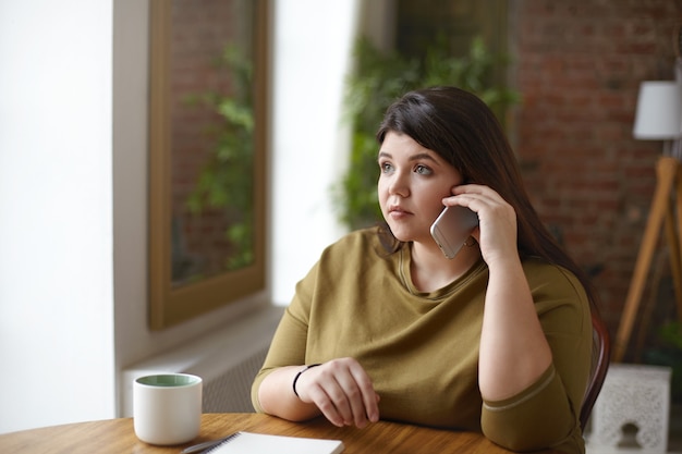 Fashionable attractive young plus size woman speaking on cell phone while drinking coffee at cafeteria with copybook lying on table in front of her. People, technology, lifestyle and communication
