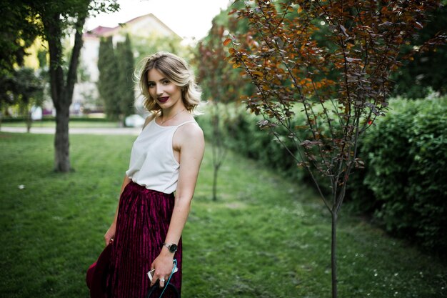Модная и красивая блондинка-модель в стильной красной бархатной велюровой юбке, белой блузке и шляпе позирует в парке с телефоном и наушниками