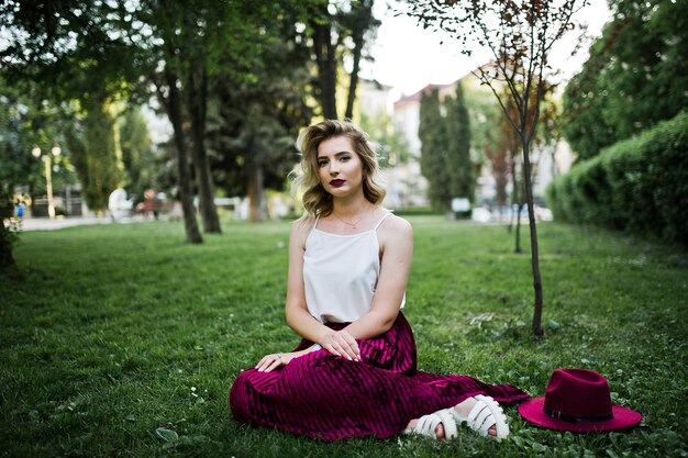 Модная и красивая блондинка-модель в стильной красной бархатной велюровой юбке, белой блузке и шляпе сидит на зеленой траве в парке