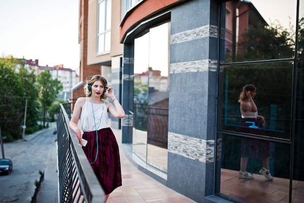 Fashionable and beautiful blonde model girl in stylish red velvet velour skirt white blouse posed with phone and earphones against windows of city building