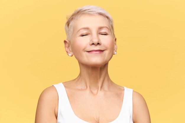 Fashionable beautiful retired Caucasian woman with pixie hairdo wearing casual clothes posing keeping eyes closed and smiling with pleasure and enjoyment, listening to good music or dreaming