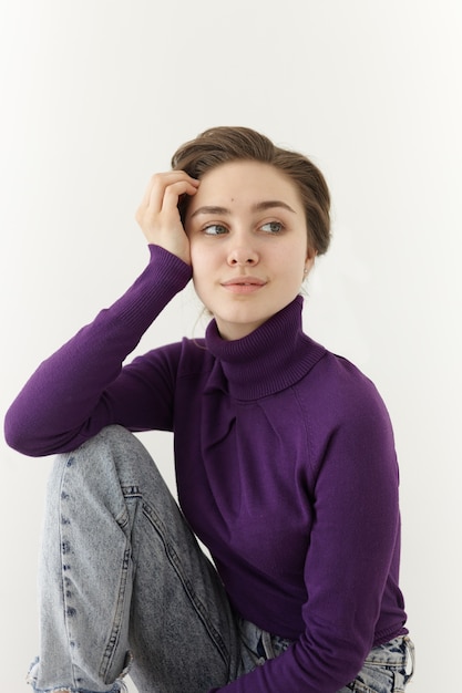 Free photo fashionable beautiful young female model wearing violet turtle neck long sleeved top and baggy jeans posing at white wall, leaning elbow on her knee and looking sideways