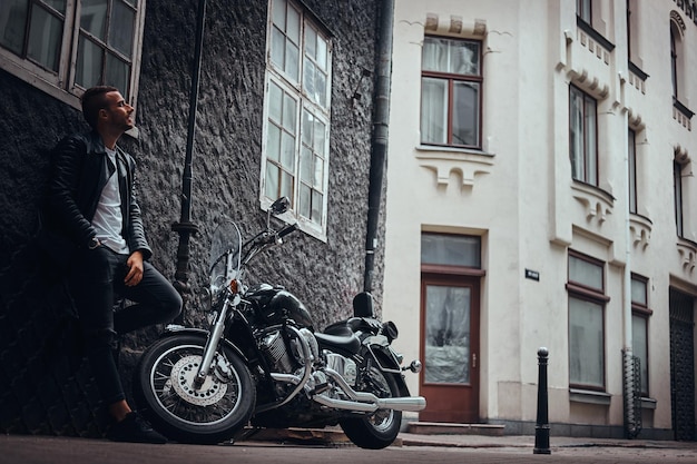 無料写真 古いヨーロッパの通りの彼のレトロなオートバイの近くの壁にもたれて黒い革のジャケットとジーンズに身を包んだファッショナブルなバイカー。