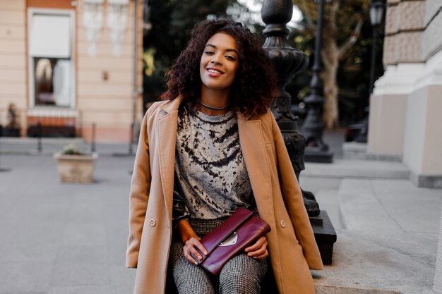 Fashionable black girl in amazing grey velvet sweater, beige wool coat, luxury jewelry accessories walking in Paris near theatre.