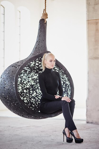 Fashionable blond female sits on a round chair in a big empty room.