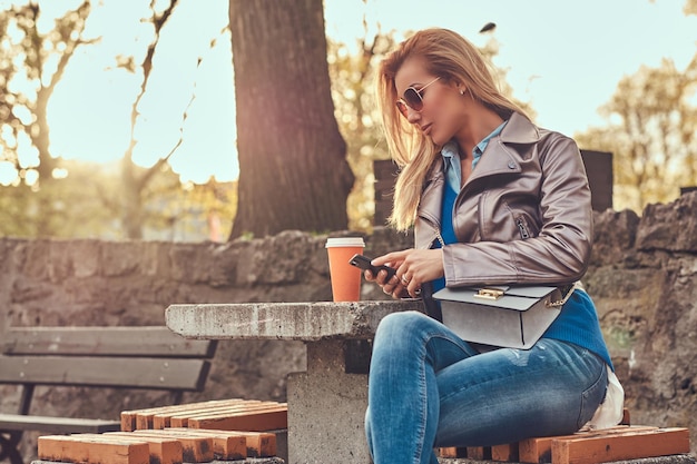 La donna bionda alla moda blogger si rilassa all'aperto, utilizzando uno smartphone mentre si siede sulla panchina nel parco cittadino contro una luce solare intensa.