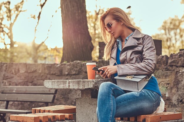 Fashionable blonde blogger female relaxes outdoor, using a smartphone while sitting on the bench in city park against a bright sunlight.